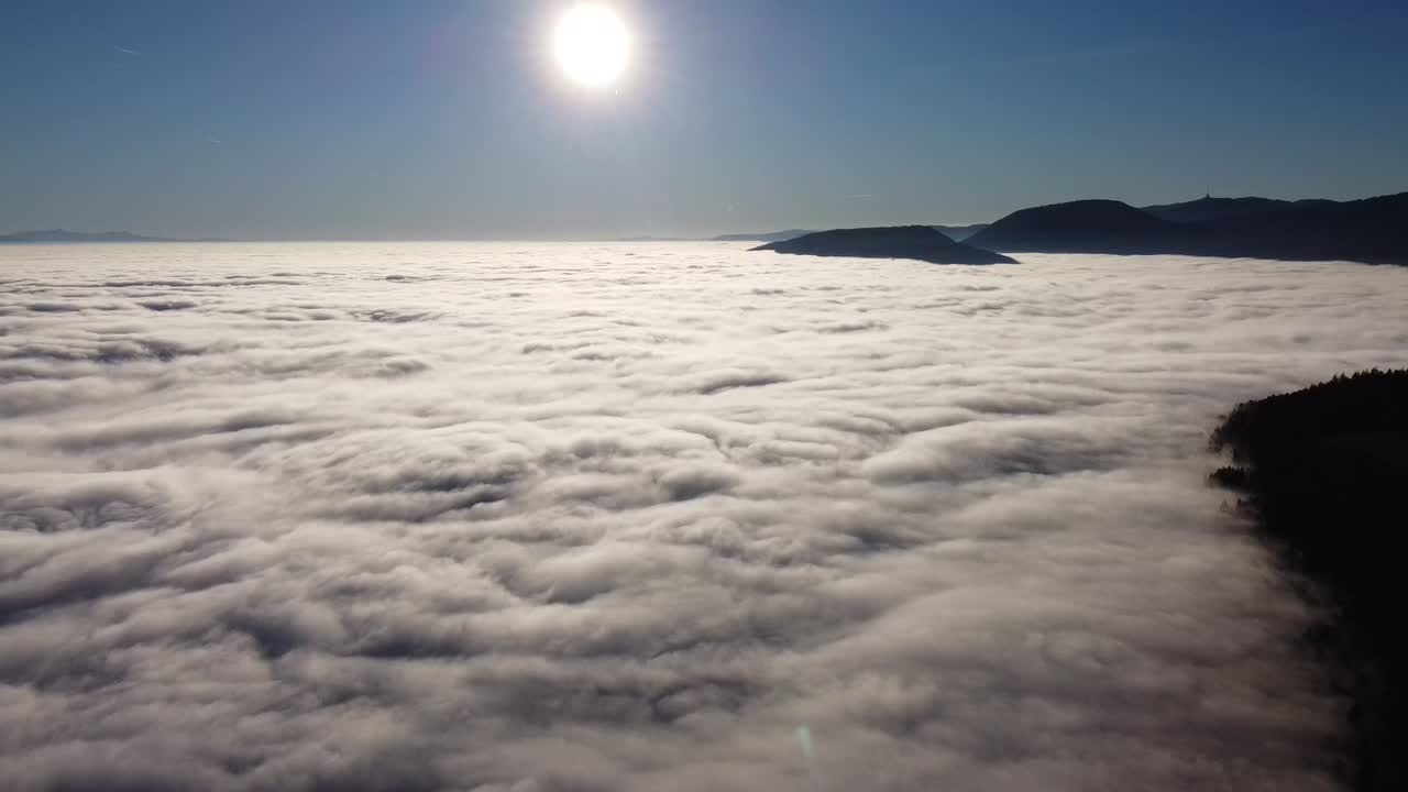 Drone flight against the sun with a bright blue sky, camera slowly tilts towards the sea of ​​fog below, in switzerland