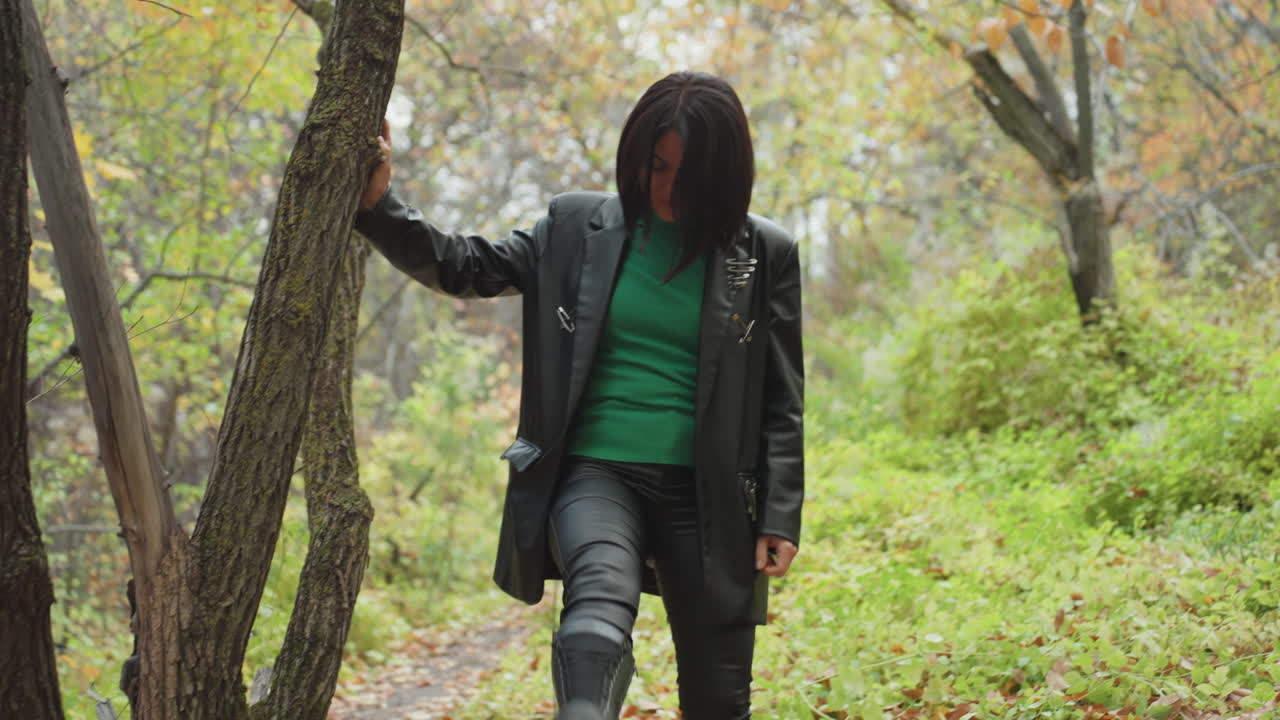 black woman in leather jacket walking through autumn woods, leaning on tree, stepping over leaf strewn path, kicking fallen leaves, wearing green sweater and boots, dreadlocks framing face