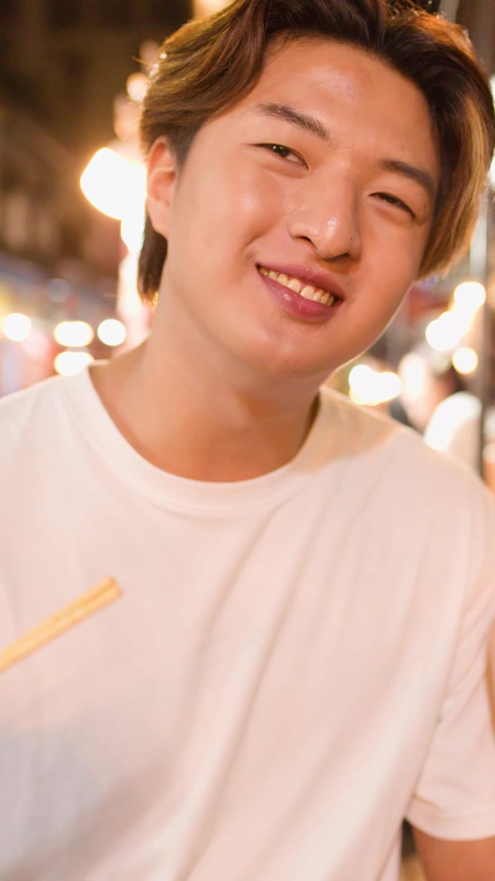 屋外のナイトマーケットで、暖色系の照明の下、男性が箸で麺を食べるクローズアップショット。
