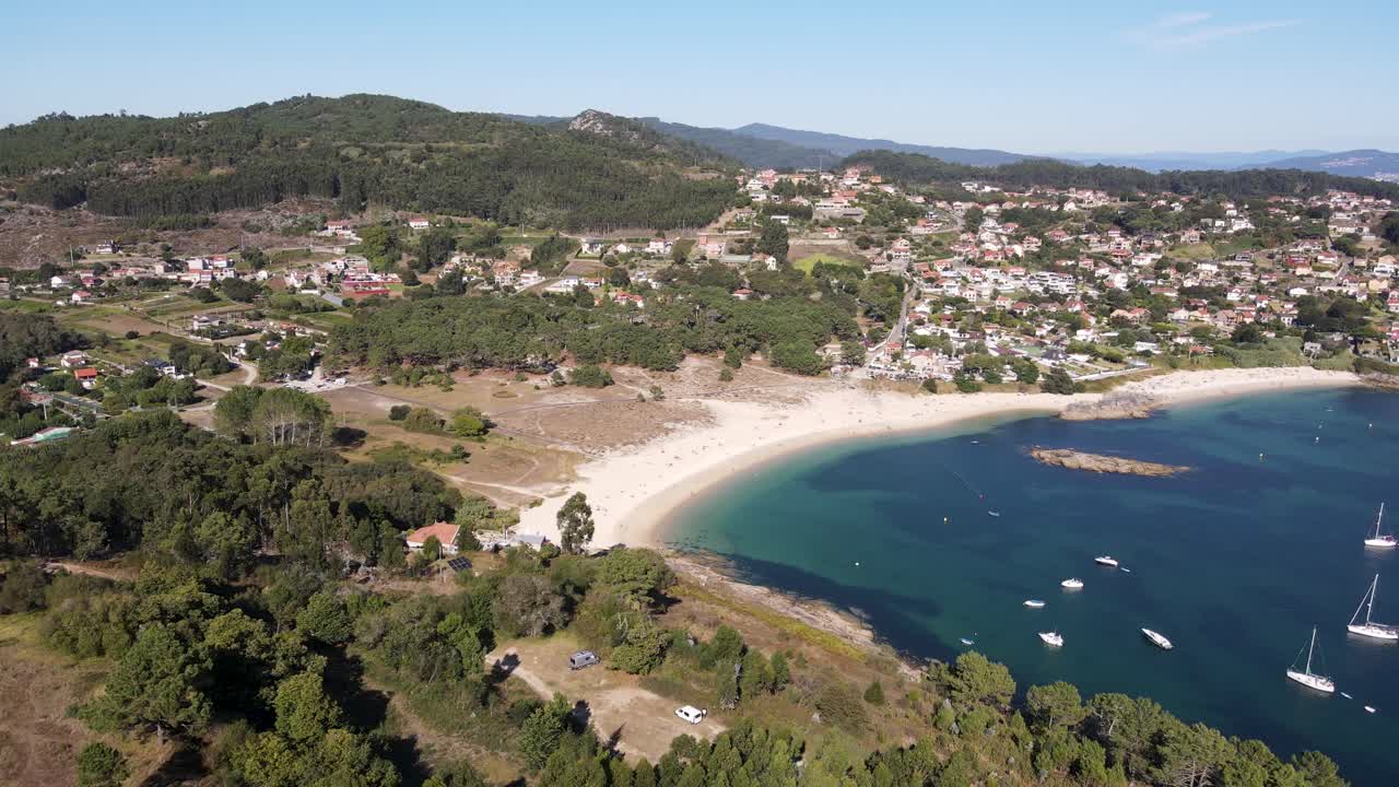Drone footage of Liméns Beach (Galicia) featuring a tranquil cove with clear blue water. The beach is framed by lush forest and hills, with scattered housing dotting the surrounding countryside