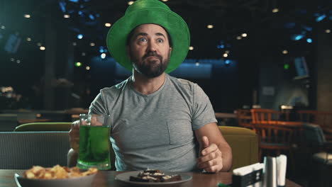 celebrar el día de san patricio en un pub. hombre con sombrero irlandés bebiendo una taza de cerveza verde y viendo un partido de fútbol en la televisión, reaccionando emocionado.