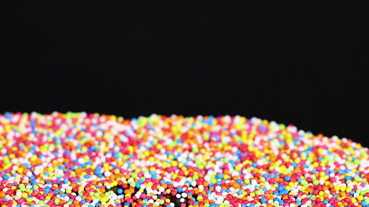 Colorful sprinkles accumulate in layers against a dark background, creating a lively and textured scene.