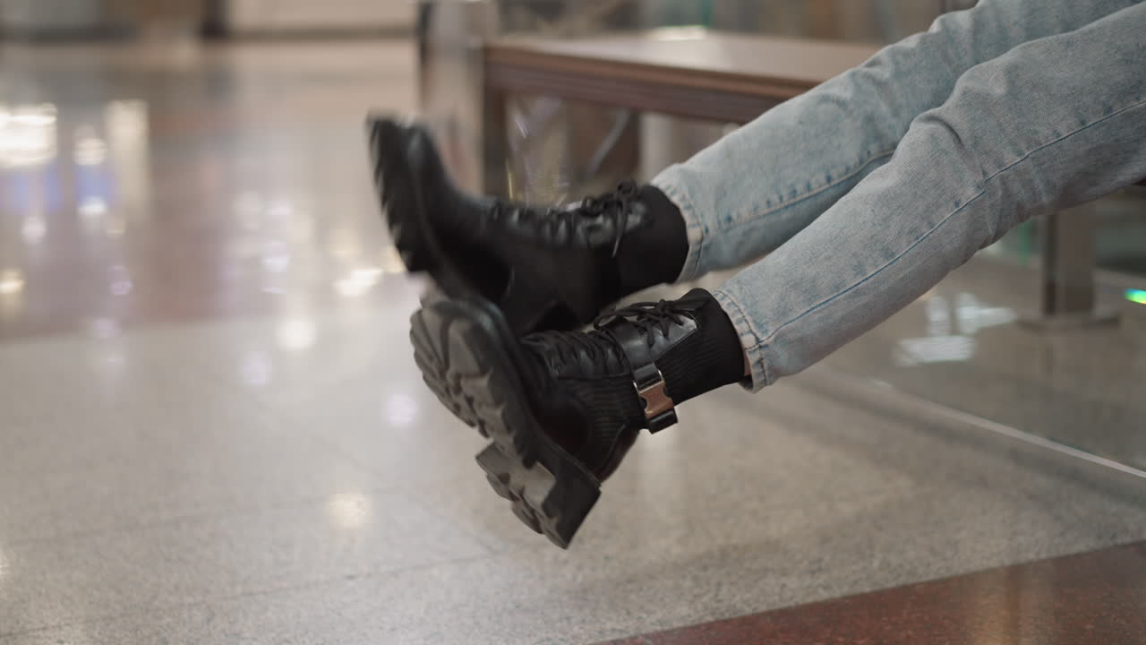 lower view of shopper swinging legs while seated on mall bench, wearing denim jeans and chunky black boots, gently kicking polished tile floor beneath glass railing with blurred holiday lights behind