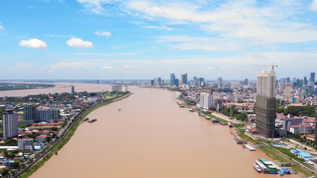 Drone video captures panoramic view of Phnom Penh's skyline. Tall buildings rise above the riverside under a vibrant blue sky, showcasing urban development and city life.