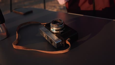 static close-up of a vintage leather-strapped camera on a table, picked up by a woman's hand.