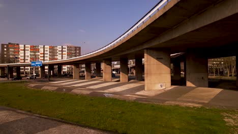 Antena-Lenta-Lateralmente-Debajo-De-Un-Gran-Paso-Elevado-De-Hormigón-Con-Gruesas-Columnas-De-Intersección-De-Carreteras-En-Un-Entorno-Urbano