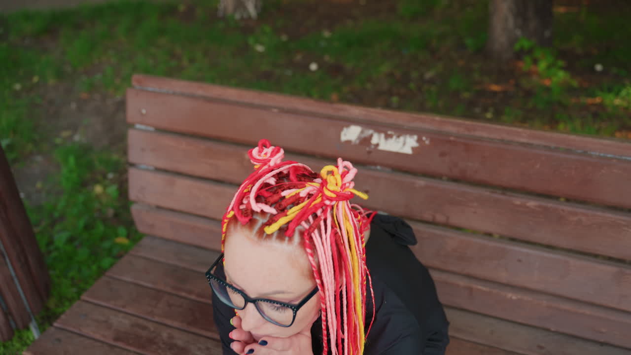 텅 빈 벤치에 앉아 화려한 드레드록을 한 여성을 내려다보는 시점, 공원 바닥과 쓰레기통이 보임, 구부정한 자세, 사색에 잠긴 듯한 멈춤, 친밀한 항공 시점, 늦은 아침 햇살