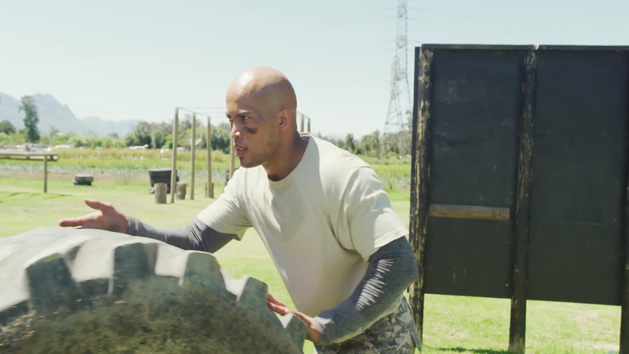 아프리카계 미국인 남성 군인이 태양 아래 군대의 장애물 코스에서 트랙터 타이어를 롤링하고 있습니다.