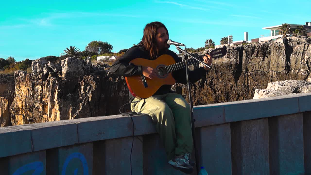 Smiling street musician sings and plays acoustic guitar at Boca do Inferno cliff viewpoint in Portugal with dramatic coastline, blue sky and cinematic slow motion movement