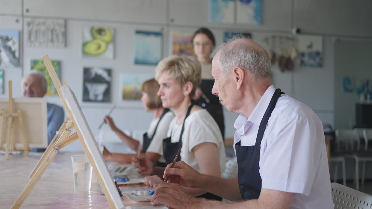한 여성 교사 는 은퇴 한 남자 에게 노인 들 을 위한 강의 에서 페인트 와 브러시 로 그림 을 그리는 방법 을 알려 준다. 한 고위 남자 는 은퇴자 들 의 집단 에게 그림 을 그린다