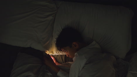 Young-Boy-In-Bedroom-At-Home-Lying-In-Bed-Using-Mobile-Phone-To-Text-Message-At-Night