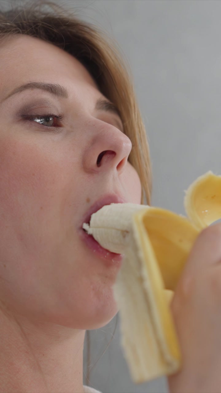 Close up of woman eating banana focusing on her mouth and fruit. sequence shows gradual consumption with visible texture details like peel separation or bite marks. Background is plain gray