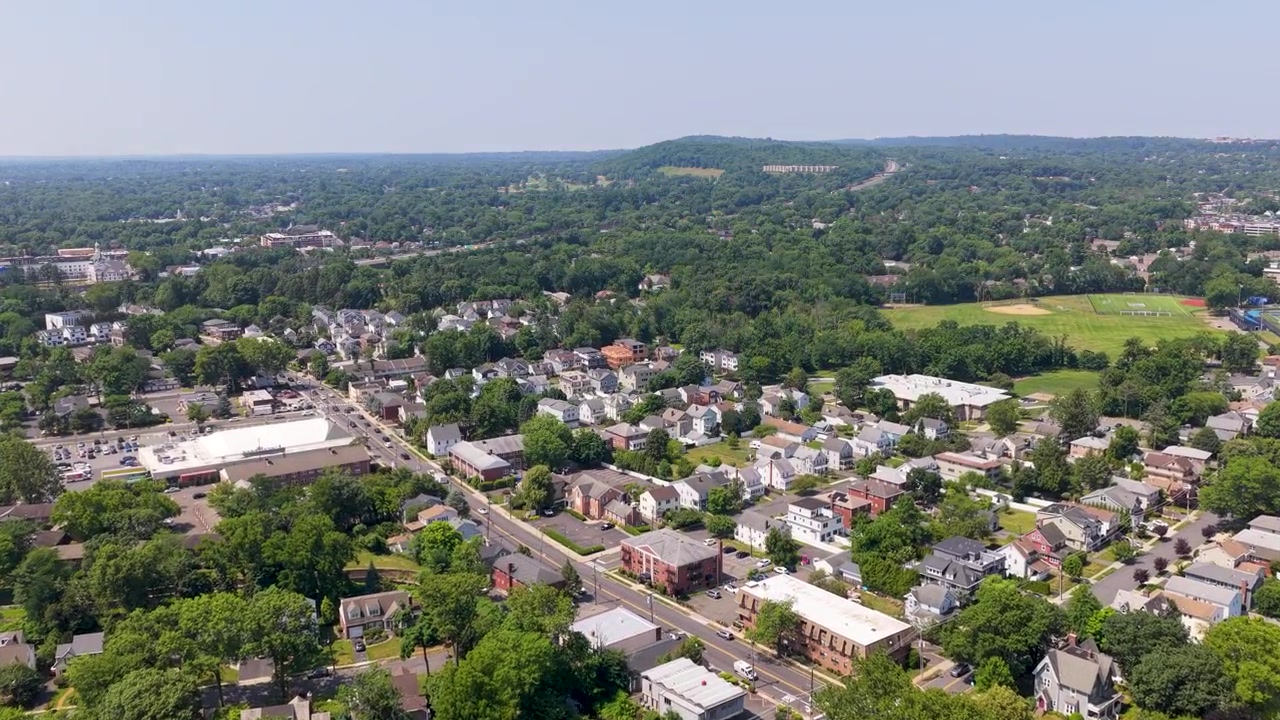 Vista aerea soleggiata con drone che sorvola la città suburbana di Milburn, New Jersey, mostrando case residenziali, alberi verdi e strade di quartiere in una bella giornata estiva con un cielo sereno.
