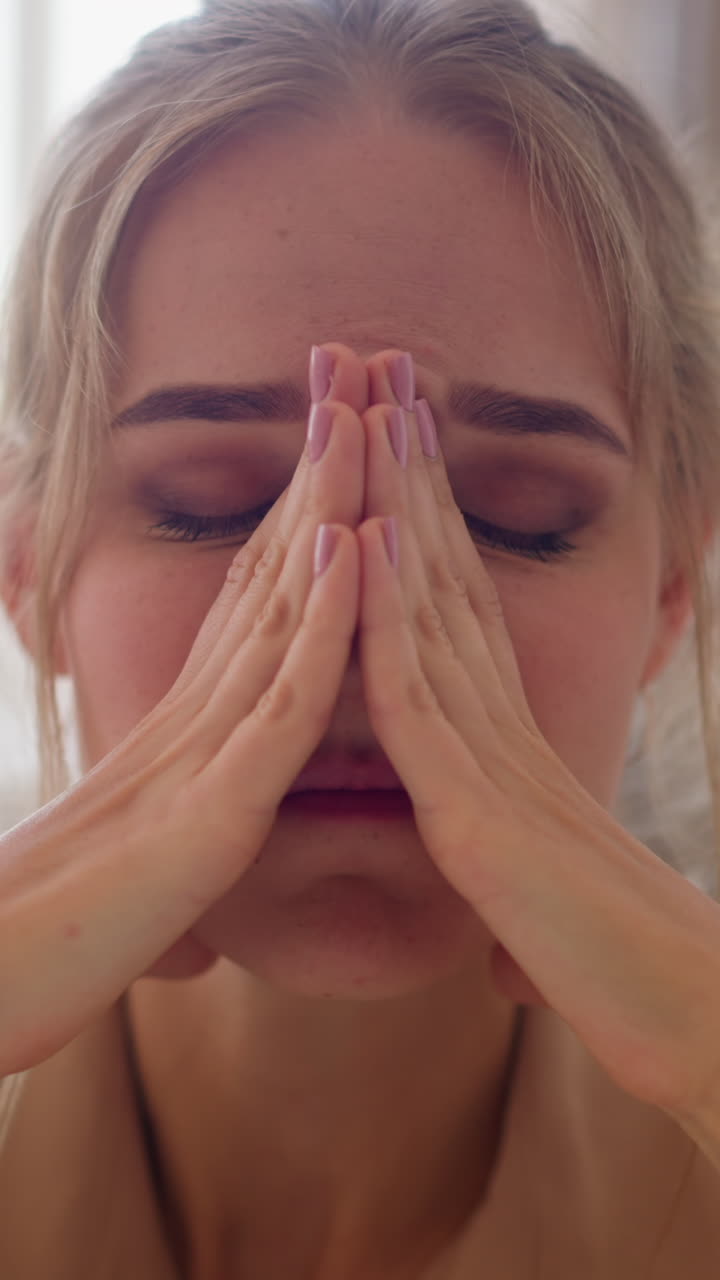Tired young woman massages weary face with hands after long time of work sitting in comfortable office against bright window closeup slow motion