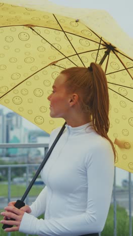 a woman under a bright yellow umbrella smiling in the rain, embracing the joy of life while overlooking a scenic view of nature in a refreshing outdoor setting