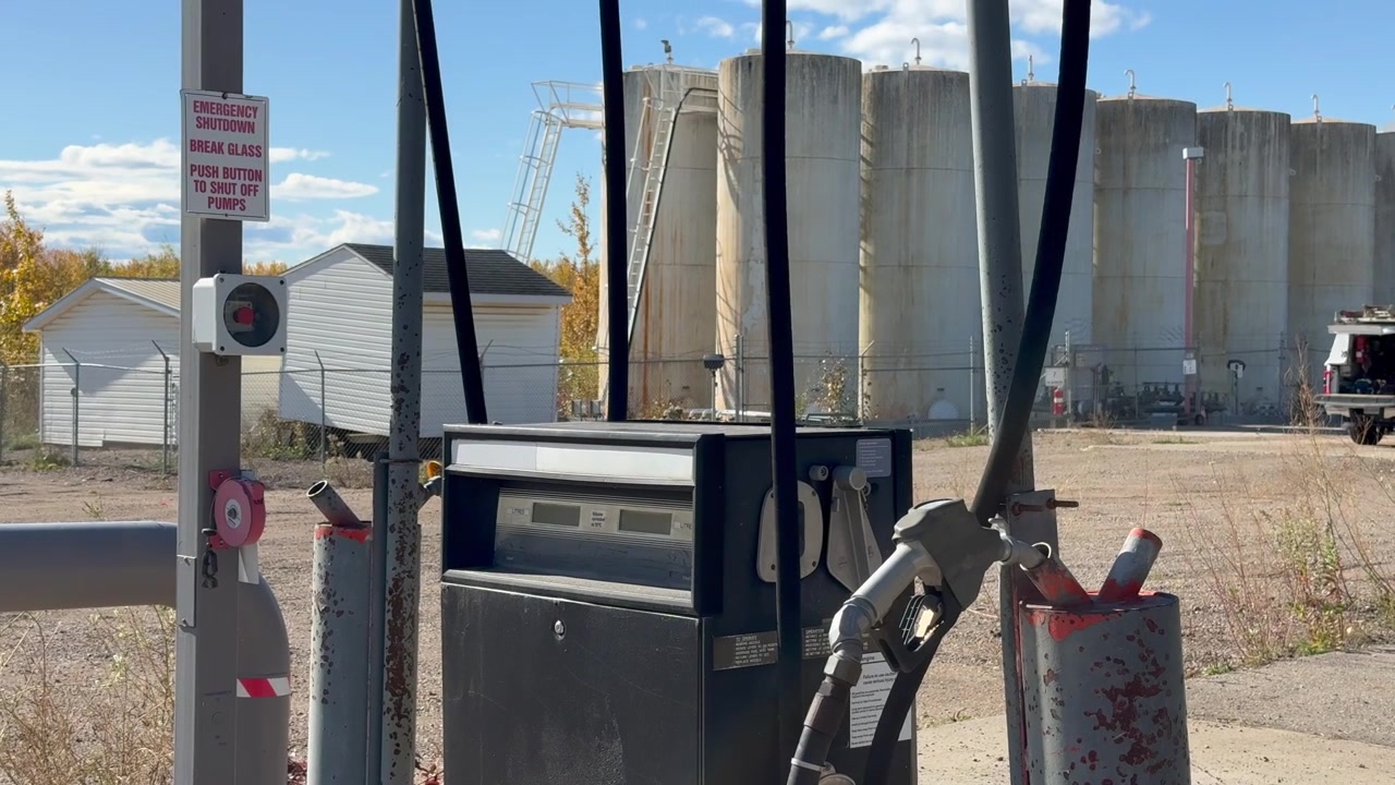 Distributore di carburante in disuso che mostra ruggine e segni di usura, con serbatoi di stoccaggio visibili sullo sfondo presso una struttura abbandonata