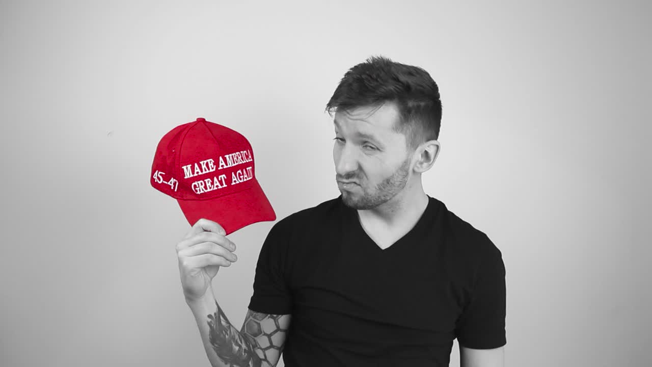Grayscale footage of a tattooed man taking off red MAGA Make America Great Again hat, looking at it unhappily and with disgust and throws it away in slow motion towards the camera, looking forward.