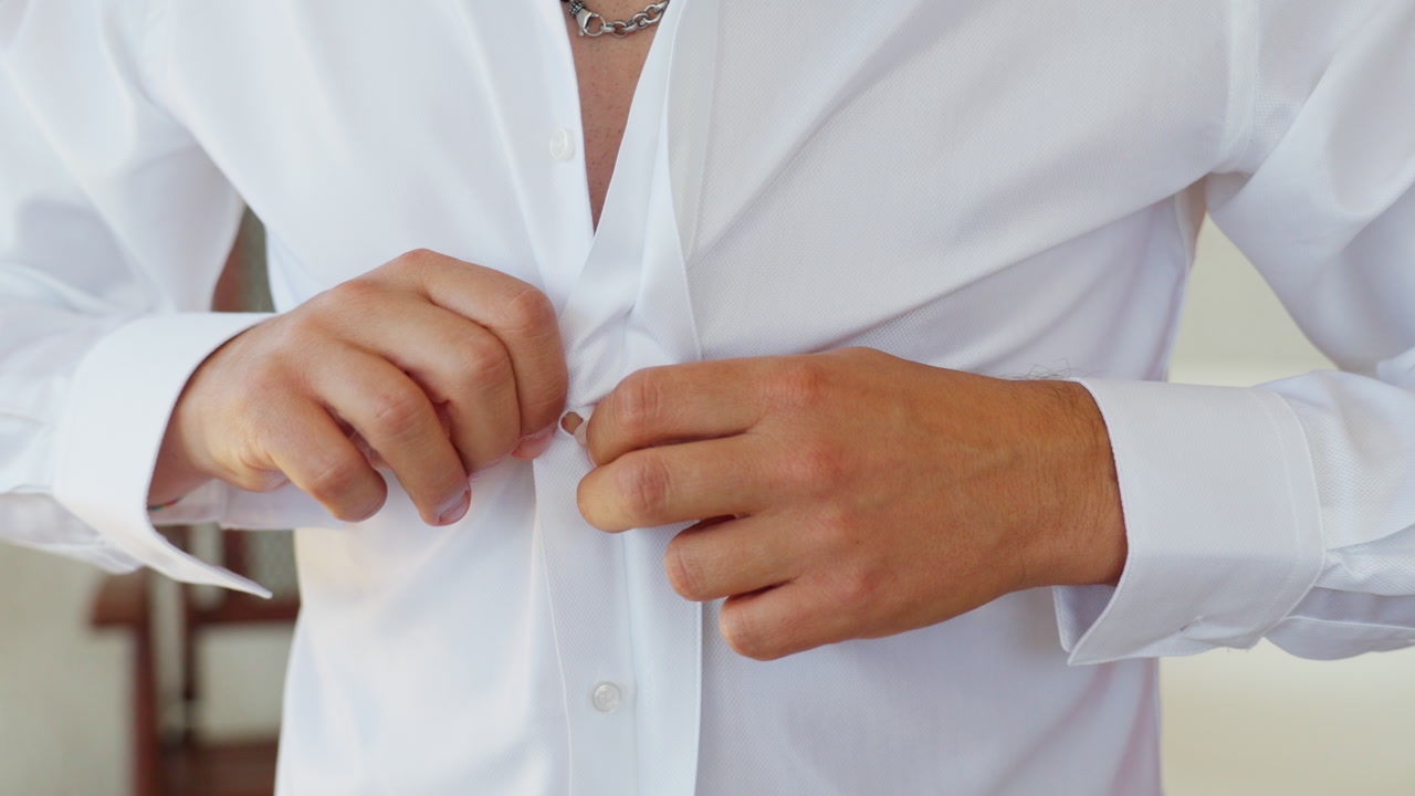 Close-up panning shot of groom buttoning white shirt during wedding preparation.