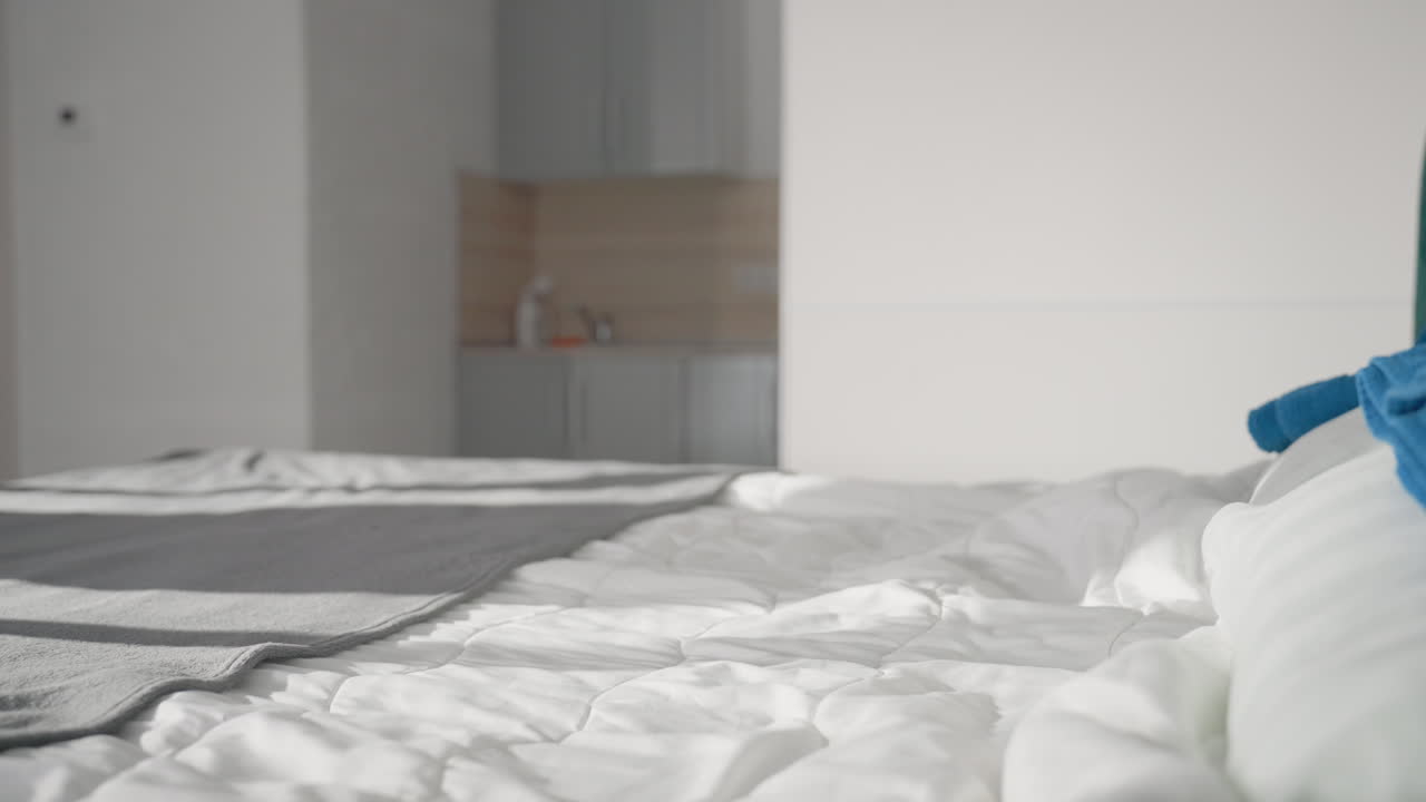 Close-up of neatly arranged bed with white quilt and grey blanket, motion blur as person tosses pillow onto bed, bright light streaming in, clean, modern bedroom interior, calm atmosphere, soft fabrics