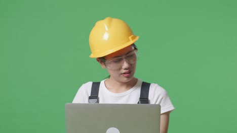 close up of asian woman worker wearing goggles and safety helmet using a laptop and having a headache while standing in the green screen background studio