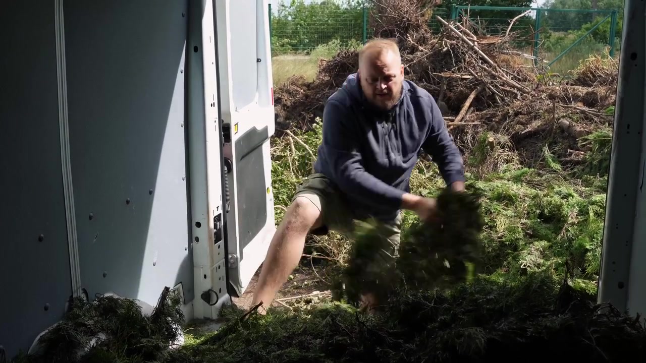 Mann i arbeidsklær trekker og kaster hauger med ferskt hageavfall fra innsiden av en varebil på et utendørs gjenvinningssted.