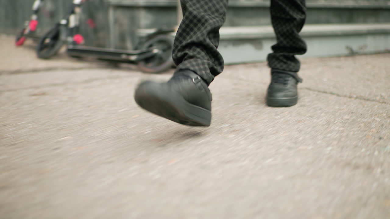 학교 건물 밖 콘크리트 계단을 내려가는 패턴 바지와 검은 신발을 신은 사람의 다리 모습, 근처에 흐릿하게 보이는 스쿠터가 주차되어 있어요