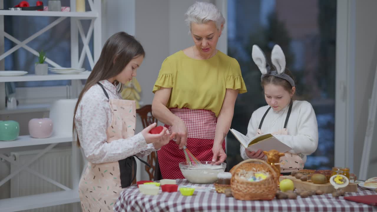 Selbstbewusste Frau mischt Backzutaten, während Mädchen ein Rezept lesen und Backformen vorbereiten. Großmutter kocht köstliches Osterdessert mit Enkelinnen, die ihr drinnen in der Küche helfen.