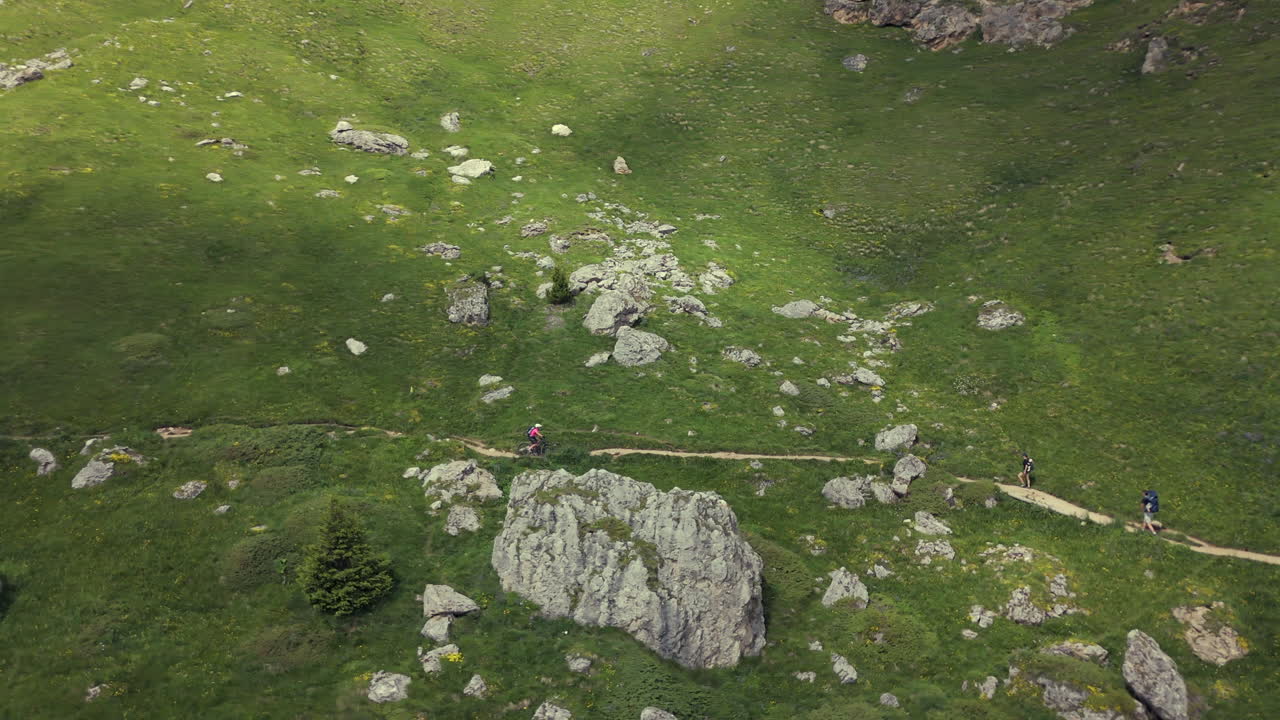 산악 자전거 타는 사람 이 산악 풍경 을 가로질러 은 바위 길 을 따라 이동 하고 있으며, 산책자 들 과 거친 지형 으로 둘러싸여 있다.