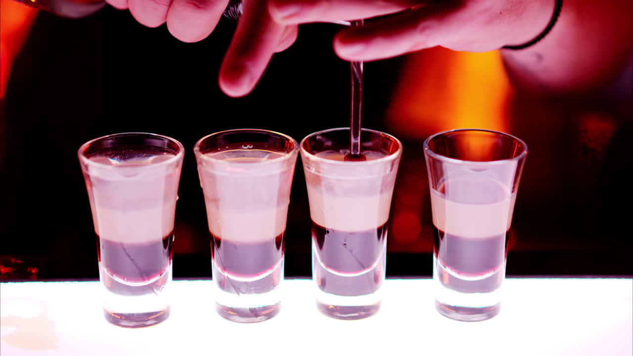 Barman making cocktail shots with liquor alcohol in three layers at the bar at night with red neon lights, slow motion