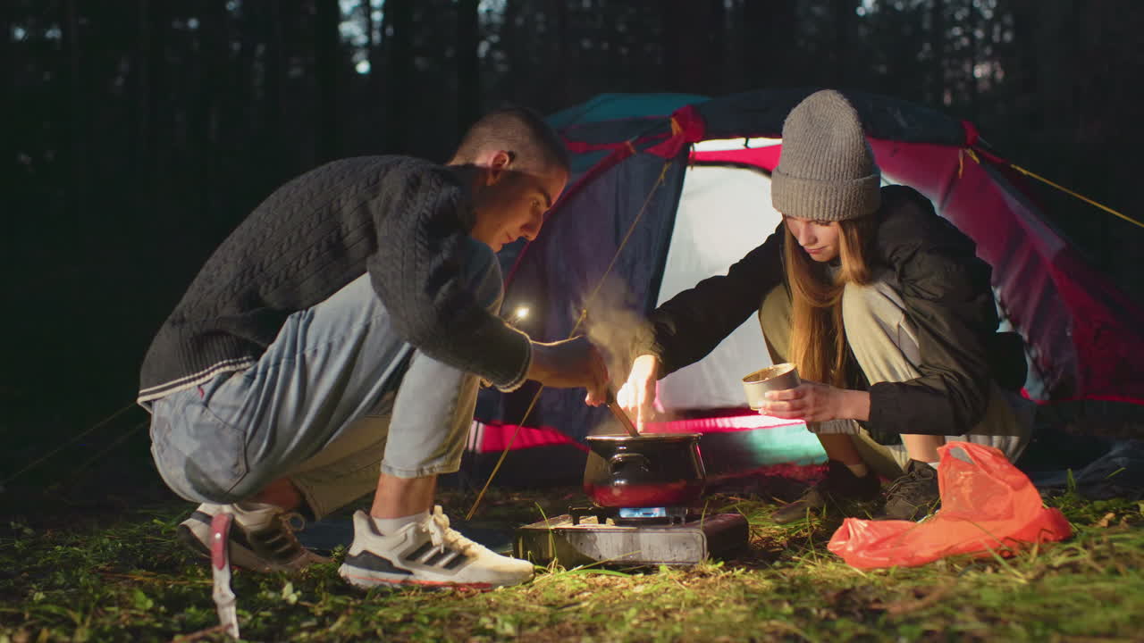 Couple camping in forest at night cooking together over gas burner near glowing tent as man stirs pot and woman holds bowl, sharing warm moment with food preparation under soft lantern light