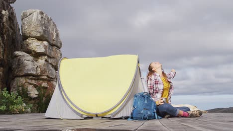 Caucasian-woman-camping,-sitting-outside-tent-with-coffee,-stretching-and-smiling-on-mountainside