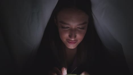 woman reading phone under a blanket at night