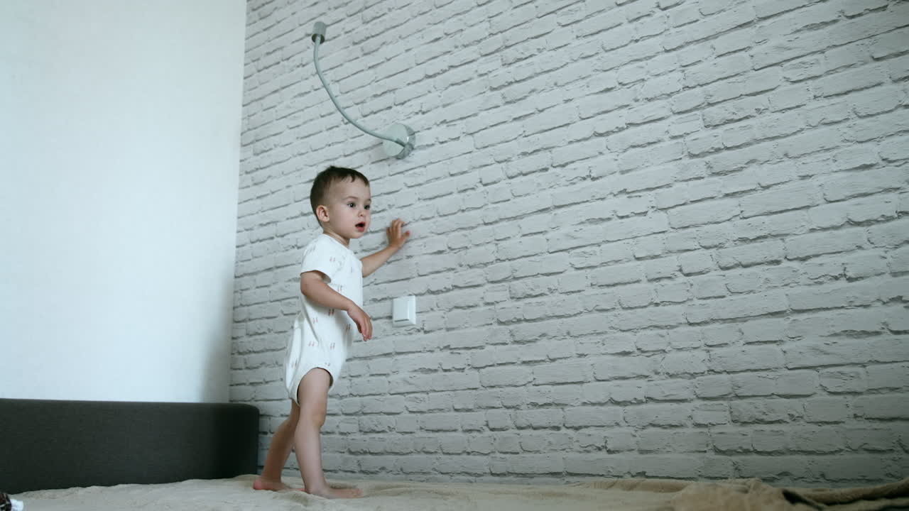 Active Caucasian toddler jumps on the bed. Baby boy switches on and off the light on the wall. Low angle view.