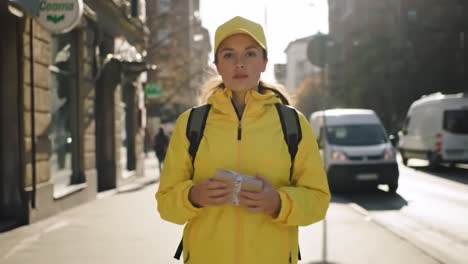 a young woman dressed in a vibrant yellow jacket and cap walks along a busy urban street. she holds a small item in her hands, exuding confidence amidst the city's hustle.