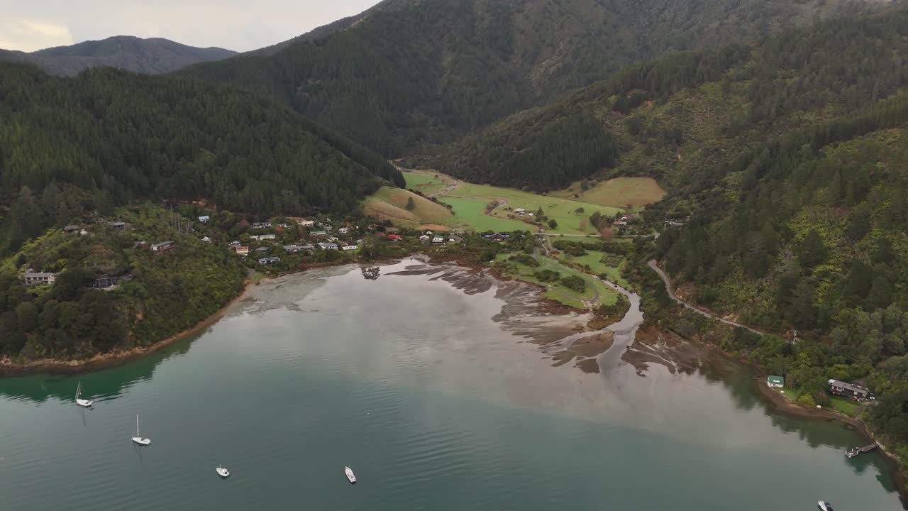 Aerial approaching shot of Whatamango Bay, with small village, fields, mountains and sailing boats on water. Top down shot. Quiet landscape in New Zealand