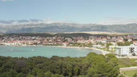 AERIAL:-Flight-over-turquoise-blue-ocean,-land-in-Novalja,-Croatia-Nature-with-houses-and-blue-sky-and-sun