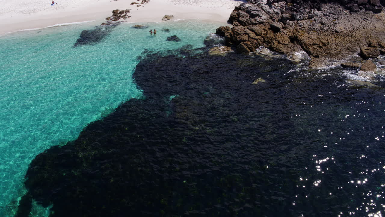 Aerial orbit of reef and rocky coastline with crashing surf and turquoise sea, friends run out of clear water