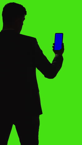 Vertical-Video-Shot-Of-Man-Holding-Blue-Screen-Mobile-Phone-Standing-Silhouetted-Against-Green-Screen-From-Behind-1