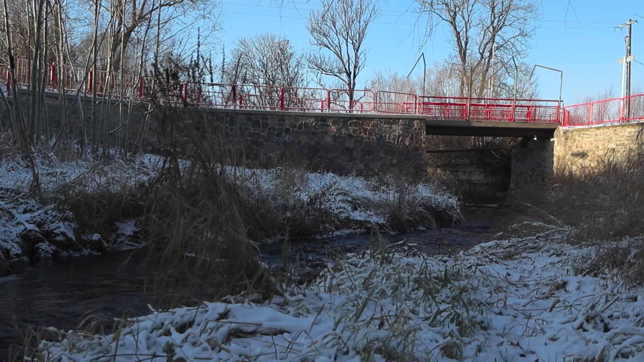 은 강물은 겨울 동안 땅 위에 눈이 쌓인 채 은 금속 레일링을 가진 돌 다리 에서 흐르며, 태양이 그 위에 빛나고 있습니다. 강은  눈과 나무로 둘러싸여 있습니다.