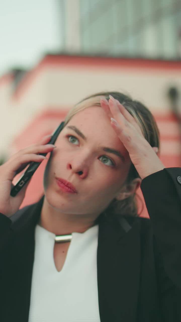 Frustrated white lady in professional outfit expressing exhaustion while on phone call in urban environment, hand raised in stress, people blurred in distant background