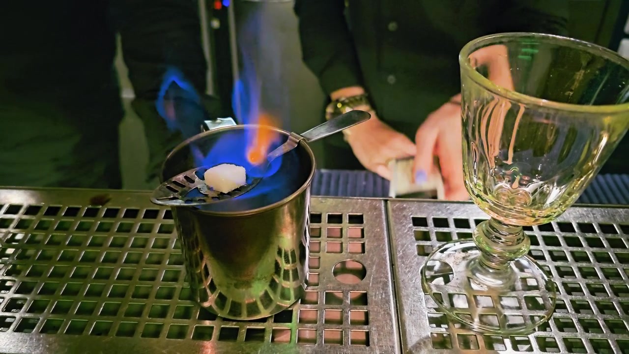 Il barista posiziona un bicchiere sul bancone durante un servizio cocktail scenografico, con fiamma blu, zolletta di zucchero, cucchiaio forato, bicchiere di metallo, vassoio raccogligocce in acciaio e atmosfera da nightclub buia all'interno.