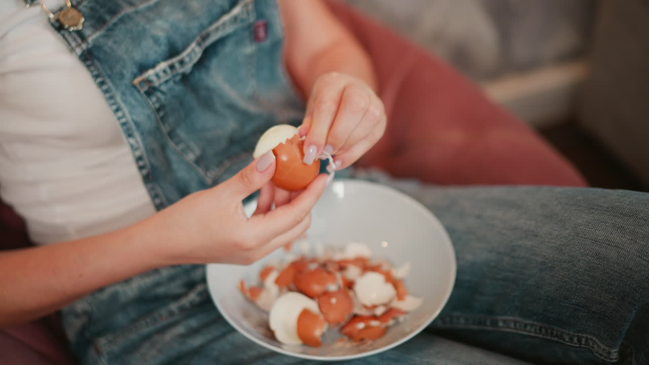 Close up of person in denim overalls carefully peeling egg shell over bowl filled with cracked shells, seated on pink beanbag with partial view of couch in background and soft lighting around
