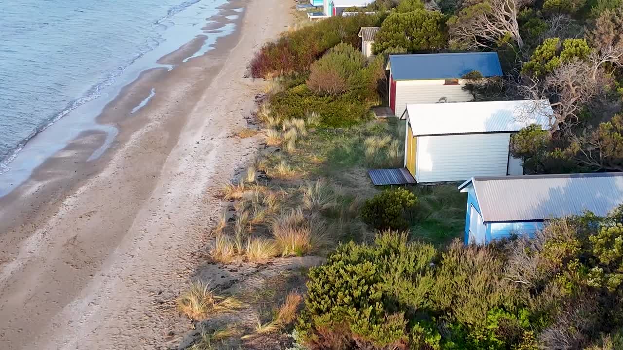 Droneoptagelser viser en person, der går tur med sin hund langs en sandstrand med farverige hytter