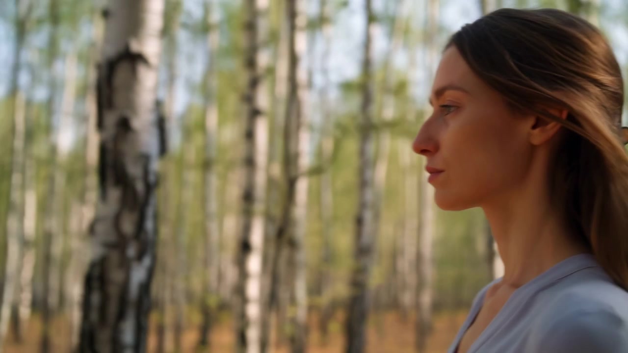 Side-view tracking composition keeps woman framed at right while birch trunks shift across softly blurred woodland, with warm sunlight illuminating her face, brown hair, and gray top.