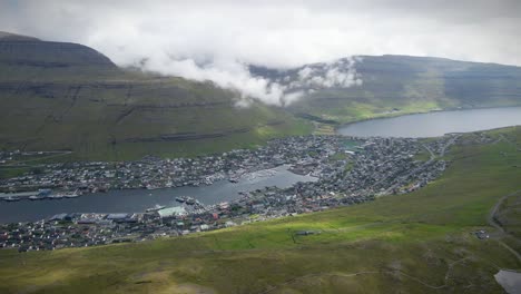 klaksvík, la segunda ciudad más grande de las islas feroe, enclavada entre las montañas y el mar, muestra el puerto con barcos, casas coloridas y nubes bajas que cubren el paisaje, estableciendo la toma