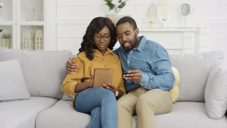 joven pareja afroamericana feliz sentada en un sofá comprando en línea con tableta y tarjeta de crédito
