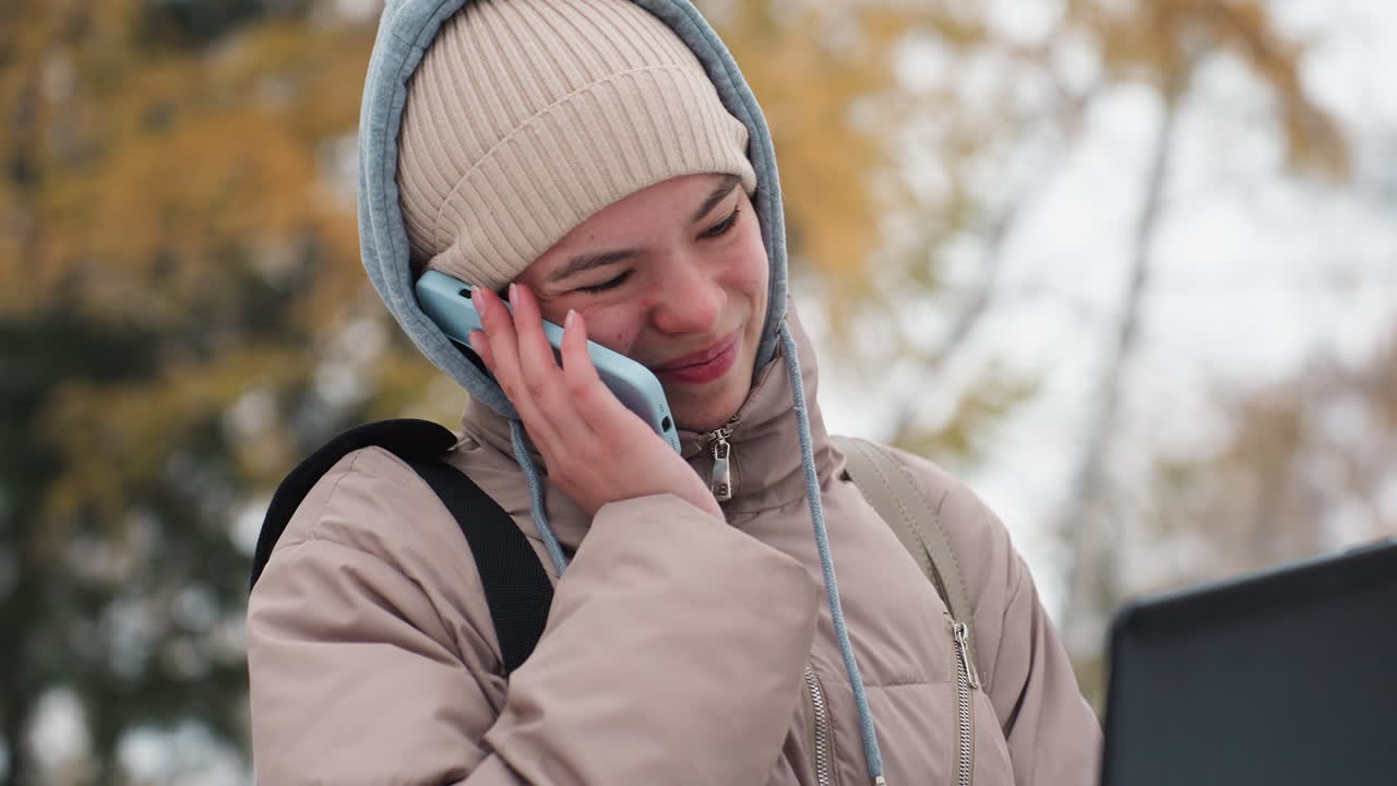 Close view of light skin lady smiling while holding phone with blue case against ear and supporting it with hand, working on laptop outdoors during cold season with soft background blur