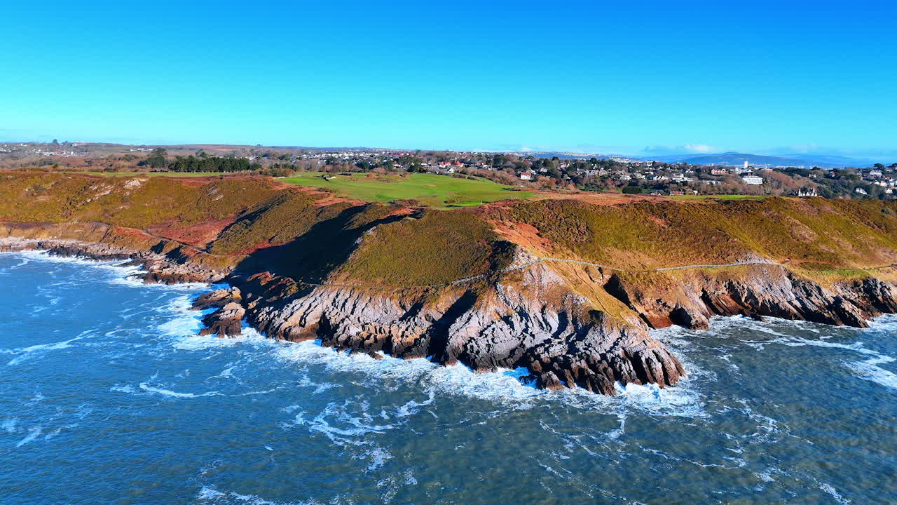 Uma costa se estende em direção à água com formações rochosas e colinas verdes ao fundo. As ondas quebram nas rochas enquanto o sol brilha intensamente no céu.