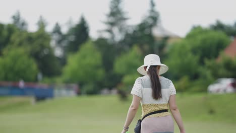 a rear view of a middle-aged woman, wearing a handmade knitted dress and a sun hat, on a stroll in a town park