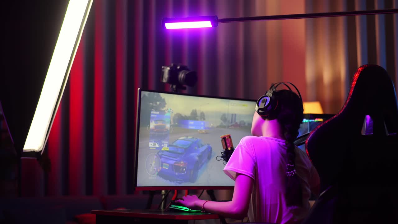 Back View Of Asian Girl Streamer Stretching While Playing Car Racing Game Over Network On Personal Computer. Live Stream Video Game, Desk Illuminated By Rgb Led Strip Light